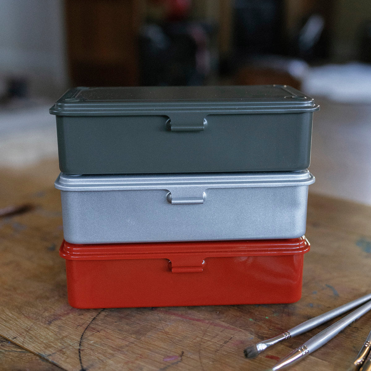 A green metal toolbox with a secured lid, placed on a wooden surface alongside stationery items like pens and a notebook, suggesting its use as a pencil case.