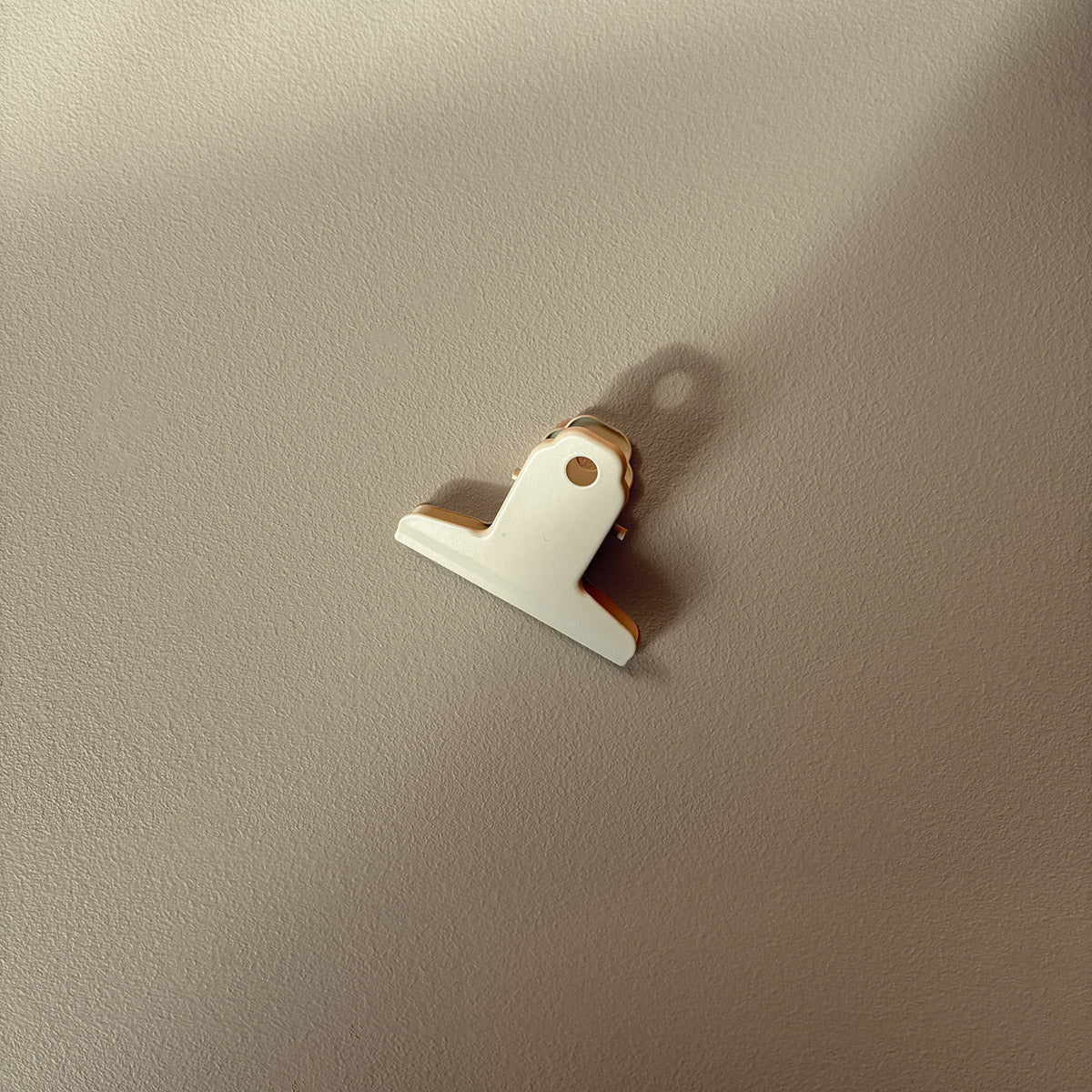 A small white metal bulldog clip with a gold-toned metal spring and backing, photographed against a textured beige wall with soft shadows.