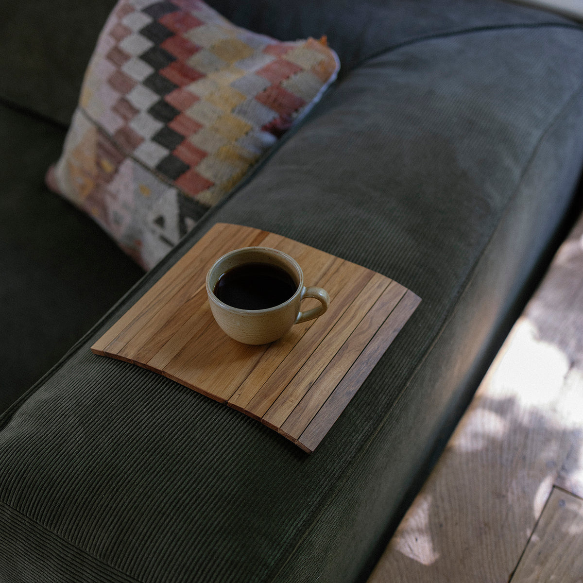 Coffee cup on a wooden couch arm tray on a dark gray couch with a patterned pillow.
