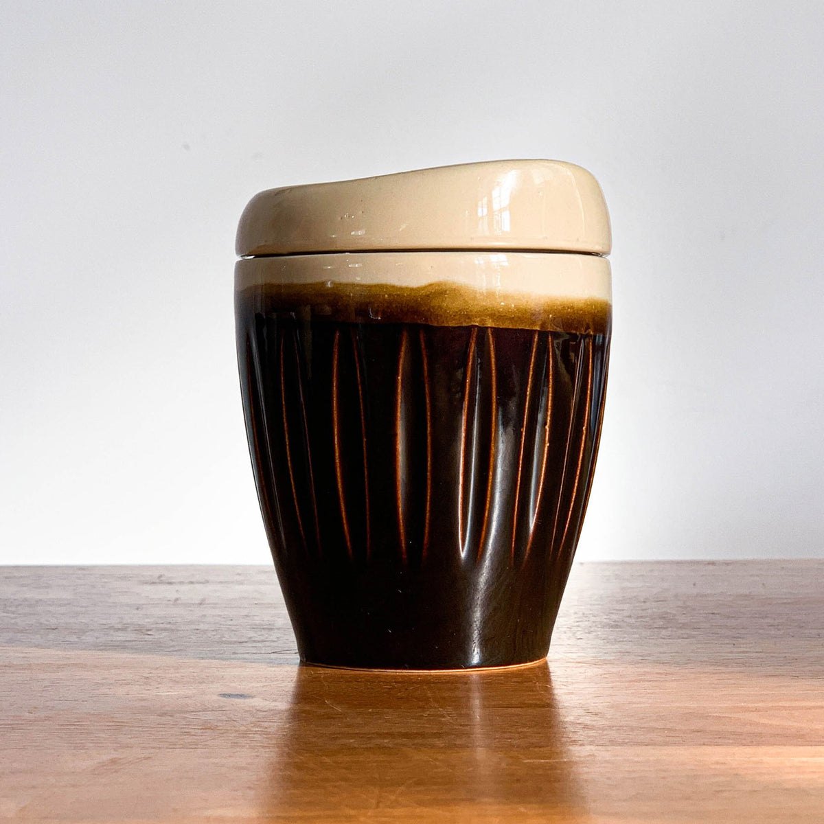 A brown ceramic reusable mug with a striped pattern and a beige ceramic lid placed on a wooden surface.