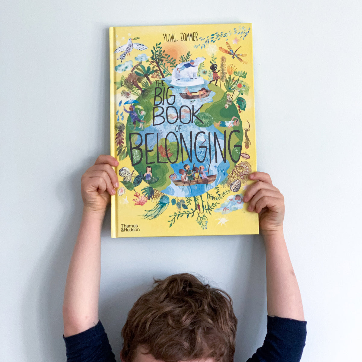 A child holding up a colorful book titled 'THE BIG BOOK OF BELONGING' with illustrations of various animals and nature.